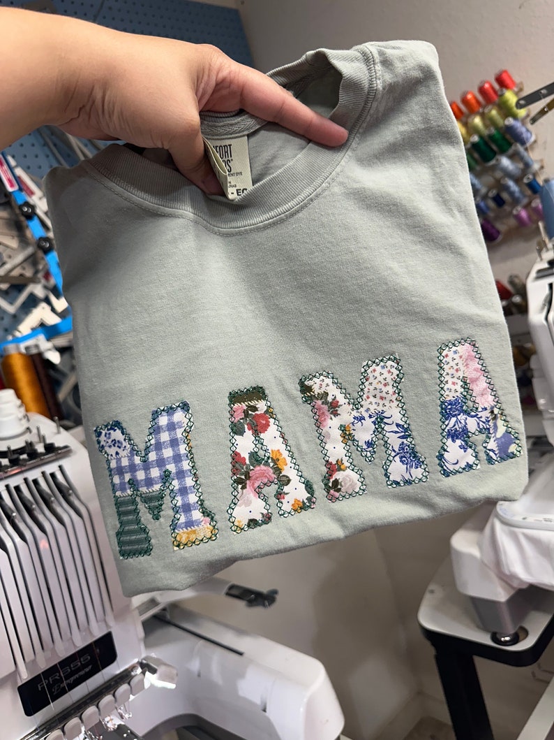 May include: A light green t-shirt with the word "MAMA" spelled out in large, colorful letters. The letters are made of different fabrics, including floral print, plaid, and solid colors.