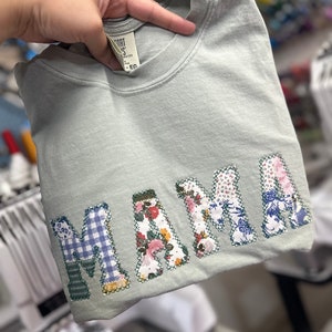 May include: A light green t-shirt with the word "MAMA" spelled out in large, colorful letters made of fabric scraps. The letters are stitched together with a white thread.
