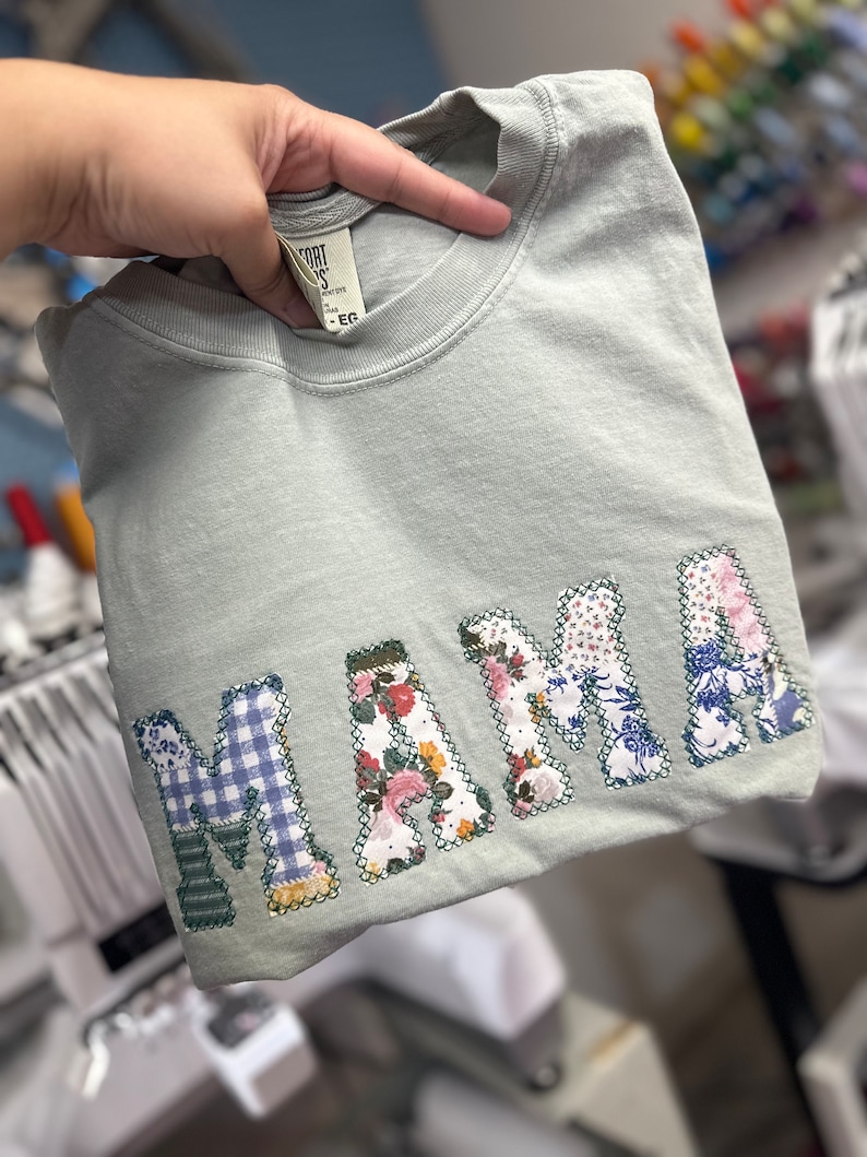 May include: A light green t-shirt with the word "MAMA" spelled out in large, colorful letters made of fabric scraps. The letters are stitched together with a white thread.