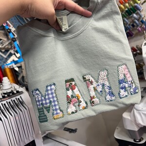 May include: A light green t-shirt with the word "MAMA" spelled out in colorful fabric patches. The patches are sewn on in a patchwork style and include floral, plaid, and geometric patterns.