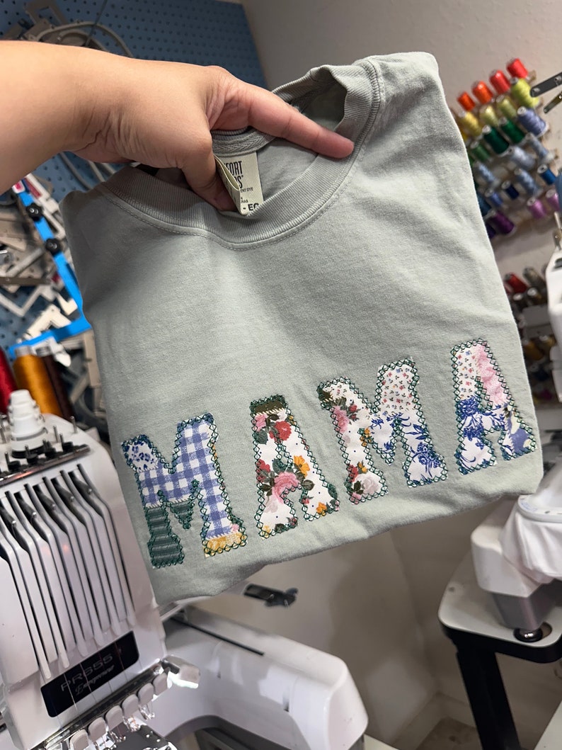 May include: A light green t-shirt with the word "MAMA" spelled out in colorful fabric patches. The patches are sewn on in a patchwork style and include floral, plaid, and geometric patterns.