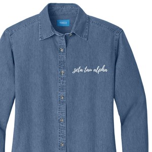 Pode incluir: Uma camisa de brim azul com botões, mangas compridas e bordado branco que diz "zeta tau alpha".