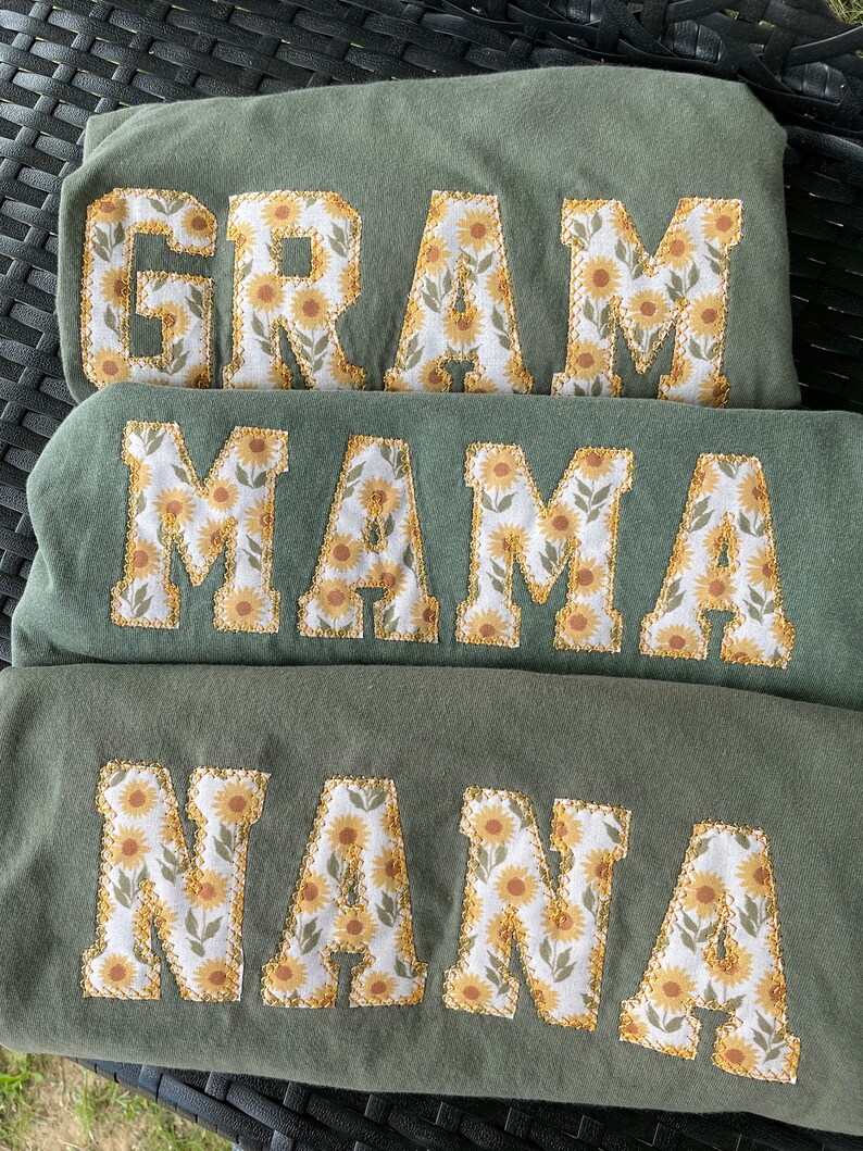 May include: Three green shirts with the words "GRAM", "MAMA", and "NANA" embroidered in white with a sunflower pattern.