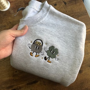 Puede incluir: Una sudadera gris con un diseño bordado de una olla de oro de dibujos animados y un trébol de cuatro hojas. La olla de oro lleva una corona y tiene una expresión feliz. El trébol también sonríe y tiene un tallo verde.