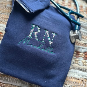 May include: A navy blue hooded sweatshirt with the letters "RN" embroidered in white with floral accents. The name "Michelle" is embroidered below the letters.