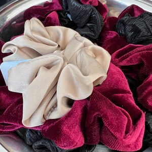 May include: A close-up of a bowl filled with a variety of hair scrunchies. The scrunchies are made of velvet and come in black, burgundy, and beige.