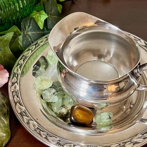 May include: A silver creamer with a decorative handle sits on a silver plate with a floral design. The plate is filled with green and brown stones.
