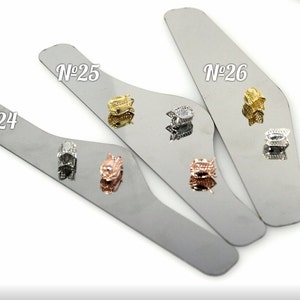 May include: Three silver metal display trays with the numbers N°24, N°25, and N°26 printed on them. Each tray has a small, decorative, metal charm with a clear stone in the center. The charms are in different colors: silver, rose gold, and gold.