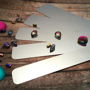 May include: A set of five silver metal rulers with a rainbow iridescent gemstone on each ruler. The rulers are arranged in a fan shape on a wooden surface. There are also several small, iridescent gemstones scattered around the rulers.
