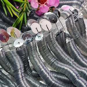 May include: Close-up of silver sequins on a string, arranged in curved lines. Pink flowers and green leaves are in the background. The sequins are round and have a reflective surface, ideal for crafting and embellishments.