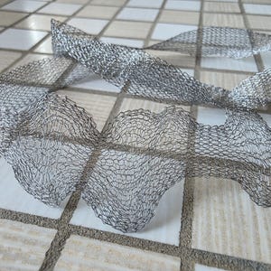 May include: A length of silver-coloured, flexible mesh ribbon. The ribbon has a delicate, open weave pattern, and is arranged in a flowing, undulating shape. The ribbon is likely used for decorative purposes, such as embellishing crafts or gifts.