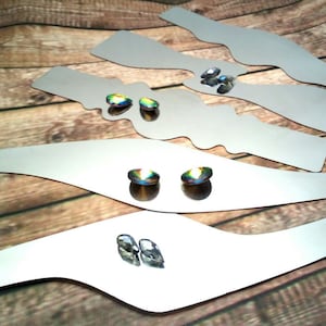 May include: Four silver-colored metal shapes, each with a curved design, are arranged on a wooden surface.  Three iridescent gemstones are placed on the shapes.