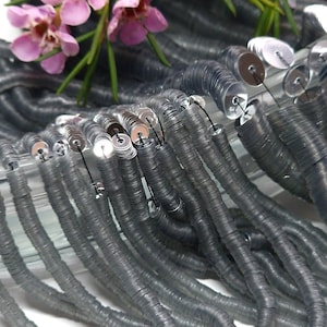 May include: A strand of silver sequins, arranged in a coil shape, with a light gray color. The sequins are small and round, with a slightly iridescent finish.