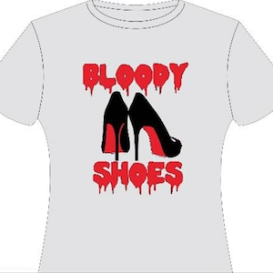 Puede incluir: Camiseta blanca con un diseño gráfico rojo que dice "BLOODY SHOES" con un efecto de goteo. El gráfico también presenta dos zapatos negros de tacón alto con suelas rojas.