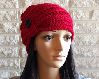 May include: A red crocheted beanie with a large button on the side. The beanie is worn by a person with long dark hair.