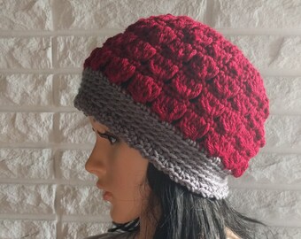 May include: A handmade, red and gray crocheted hat. The hat features a textured, scale-like pattern in red on the crown, with a gray band around the base. The hat is designed to be worn on the head and is a stylish accessory.