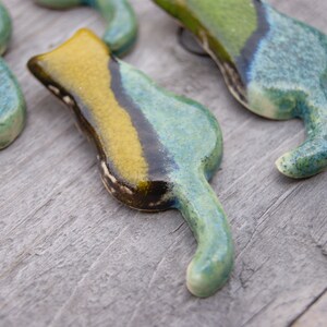May include: Four ceramic cat figurines in shades of green, blue, and yellow. The cats are stylized with long tails and are arranged in a row on a wooden surface.