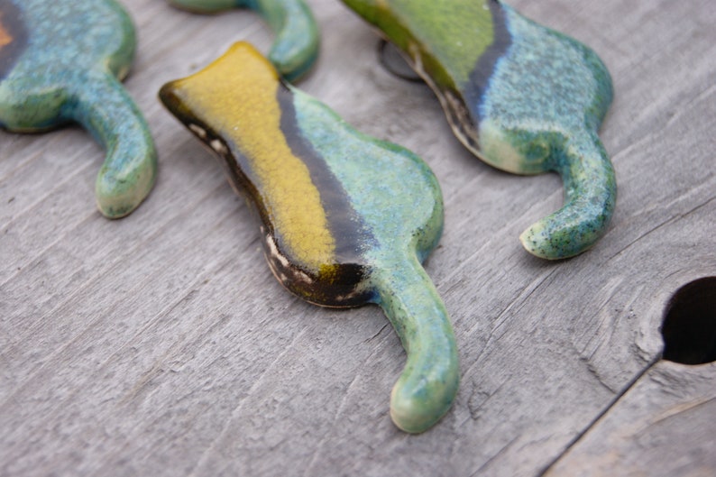 May include: Four ceramic cat figurines in shades of green, blue, and yellow. The cats are stylized with long tails and are arranged in a row on a wooden surface.