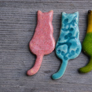 May include: Four ceramic cat figurines in different colors: pink, blue, green, and black. The cats are all facing the same direction and have a simple, stylized design.