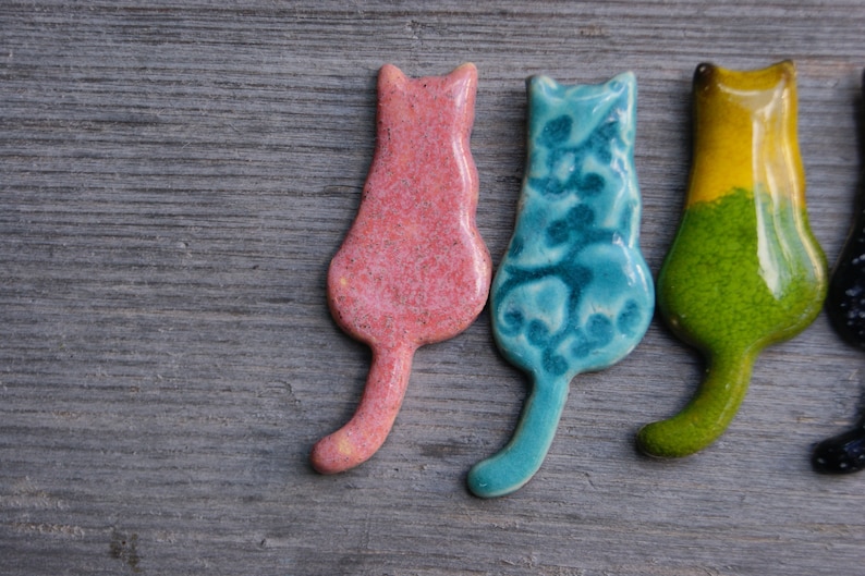 May include: Four ceramic cat figurines in different colors: pink, blue, green, and black. The cats are all facing the same direction and have a simple, stylized design.