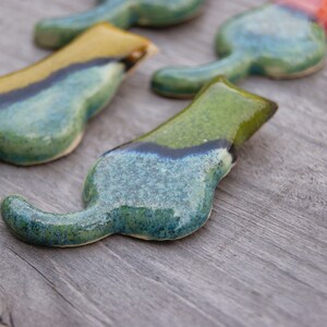 May include: Four ceramic cat figurines in various shades of blue, green, and yellow. The cats are arranged in a line, with the tails of the cats in the back of the line overlapping the heads of the cats in the front of the line.