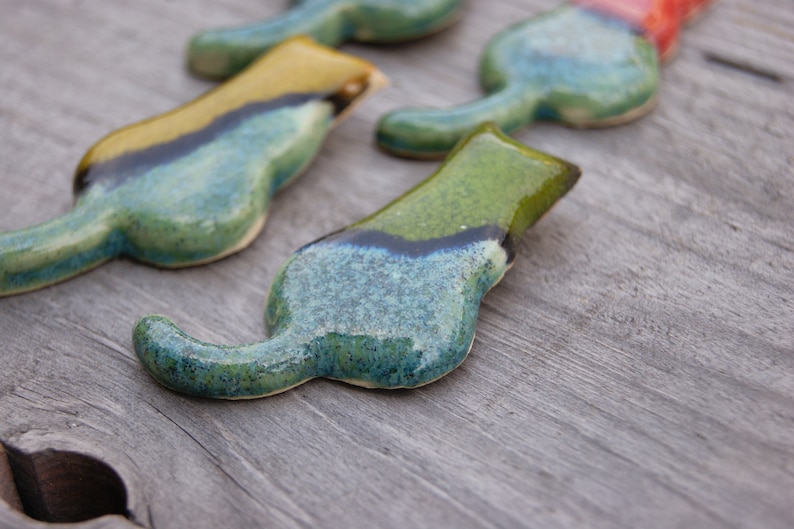 May include: Four ceramic cat figurines in various shades of blue, green, and yellow. The cats are arranged in a line, with the tails of the cats in the back of the line overlapping the heads of the cats in the front of the line.