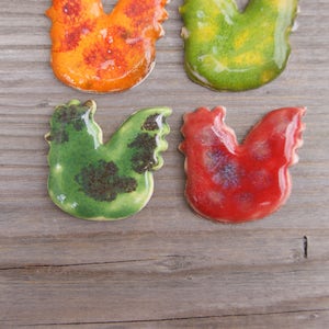 Op de afbeelding: Vier keramische haanfiguren in verschillende kleuren. De hanen zijn groen, rood, oranje en geel. Ze zijn allemaal geglazuurd en hebben een rustieke uitstraling.