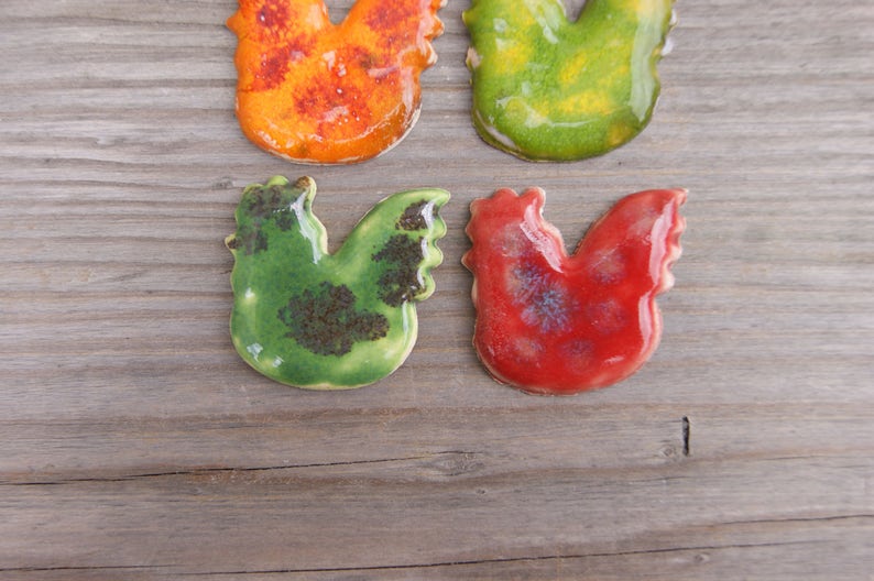 Op de afbeelding: Vier keramische haanfiguren in verschillende kleuren. De hanen zijn groen, rood, oranje en geel. Ze zijn allemaal geglazuurd en hebben een rustieke uitstraling.
