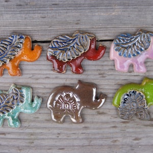 May include: Six ceramic elephant figurines with different colored glazes and intricate designs. The elephants are all facing the right and have a raised trunk. The designs include floral patterns and leaf motifs.