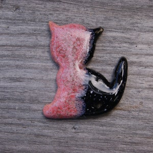 May include: A ceramic cat figurine with a pink and black glaze. The cat is sitting with its tail curled around its body.