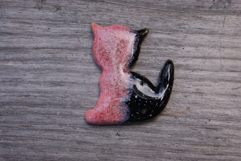 May include: A ceramic cat figurine with a pink and black glaze. The cat is sitting with its tail curled around its body.