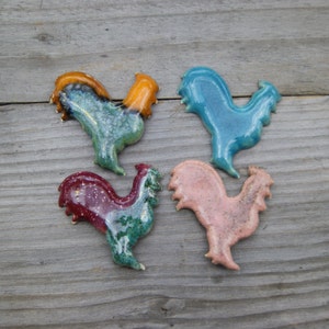 May include: Four ceramic rooster figurines in different colors: teal, orange, pink, and maroon. Each rooster has a unique glaze and design.