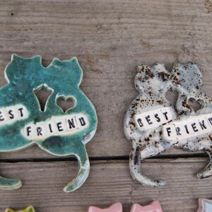 Puede incluir: Dos figuritas de cer&aacute;mica de gatos, pintadas en verde y gris con motas marrones, est&aacute;n unidas. Cada gato tiene un coraz&oacute;n recortado y una baldosa con las palabras "BEST FRIEND" en ella.