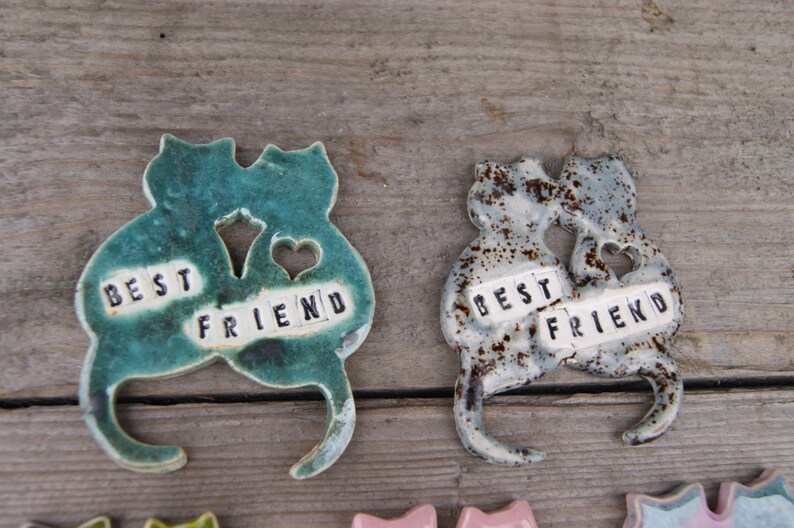 Puede incluir: Dos figuritas de cer&aacute;mica de gatos, pintadas en verde y gris con motas marrones, est&aacute;n unidas. Cada gato tiene un coraz&oacute;n recortado y una baldosa con las palabras "BEST FRIEND" en ella.
