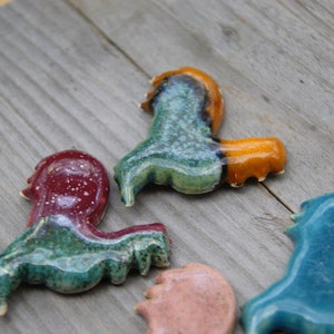 May include: Five ceramic figurines in the shape of a rooster, each with a different glaze. The roosters are in shades of blue, green, red, orange, and pink.