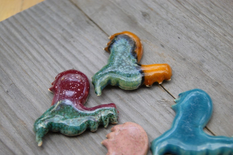 May include: Five ceramic figurines in the shape of a rooster, each with a different glaze. The roosters are in shades of blue, green, red, orange, and pink.