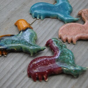 May include: Four ceramic rooster figurines in various colors, including teal, orange, green, and pink. The roosters are all in a similar pose, with their heads tilted to the side and their wings spread out.