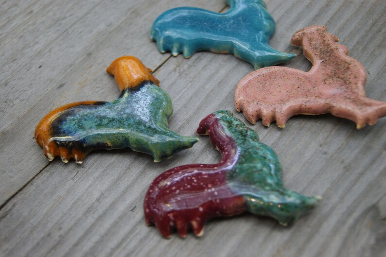May include: Four ceramic rooster figurines in various colors, including teal, orange, green, and pink. The roosters are all in a similar pose, with their heads tilted to the side and their wings spread out.