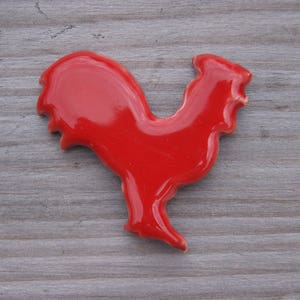 May include: A red ceramic rooster figurine. The rooster is standing on a wooden surface.