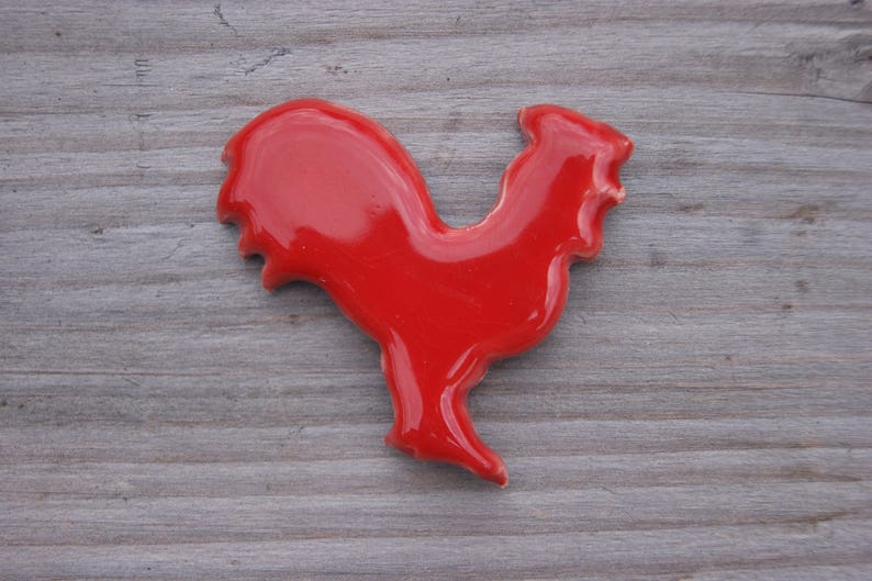 May include: A red ceramic rooster figurine. The rooster is standing on a wooden surface.