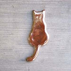 May include: A ceramic cat figurine in a brown glaze with a metallic sheen. The cat is sitting with its tail curled around its body.