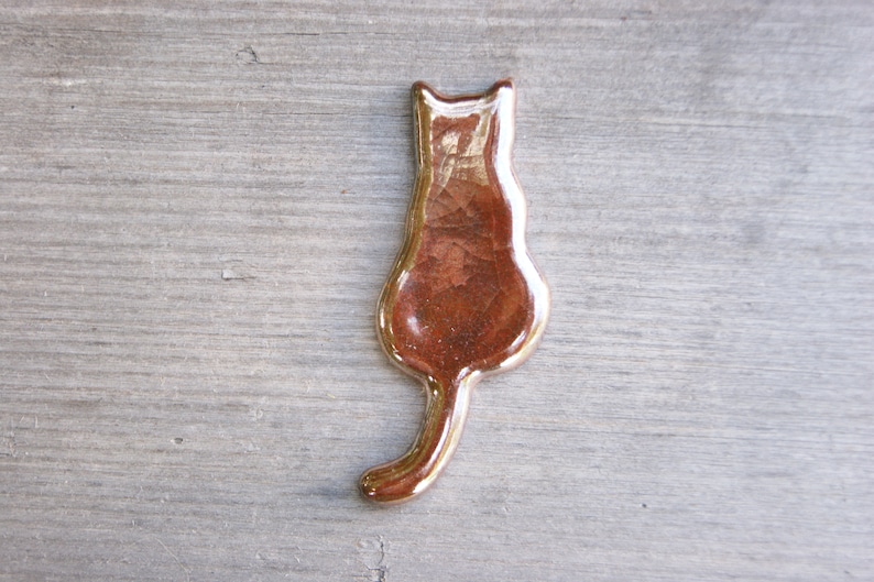 May include: A ceramic cat figurine in a brown glaze with a metallic sheen. The cat is sitting with its tail curled around its body.