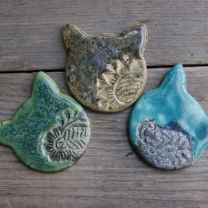 Puede incluir: Tres adornos de cer&aacute;mica con forma de cabeza de gato en verde, azul y marr&oacute;n con un acabado texturizado y esmaltado.