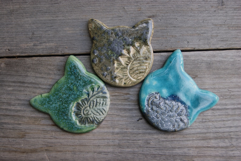 Puede incluir: Tres adornos de cer&aacute;mica con forma de cabeza de gato en verde, azul y marr&oacute;n con un acabado texturizado y esmaltado.