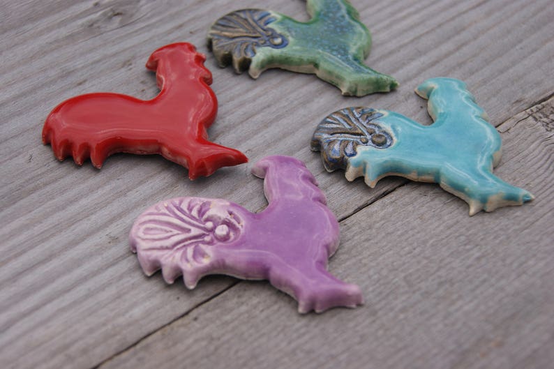 May include: Four ceramic rooster figurines in different colors: red, green, blue, and purple. Each rooster has a unique design and is a decorative piece.