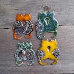 May include: Four ceramic cat figurines, each with a different glaze color: orange, green, blue, and yellow. The cats are in a cuddling pose, with intricate blue and white floral designs on their bodies.