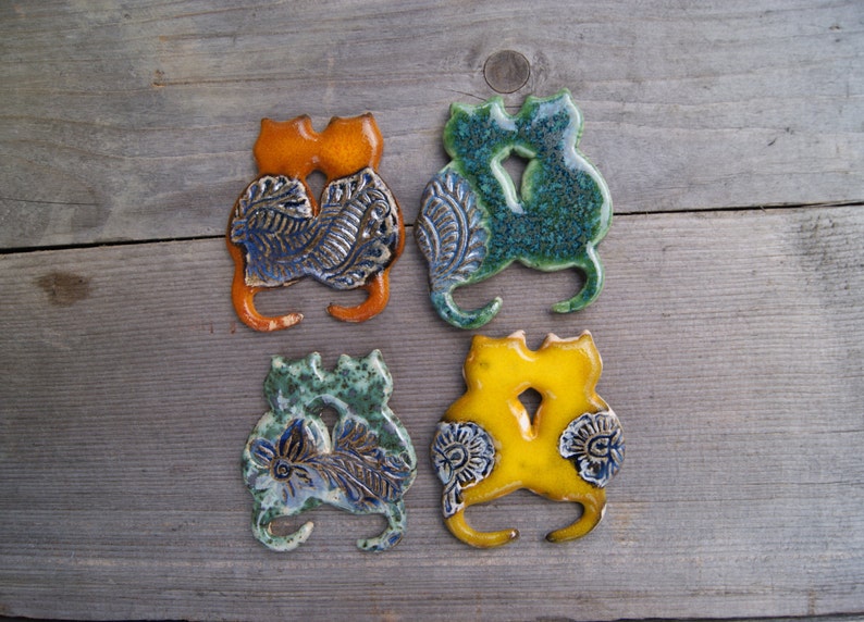 May include: Four ceramic cat figurines, each with a different glaze color: orange, green, blue, and yellow. The cats are in a cuddling pose, with intricate blue and white floral designs on their bodies.