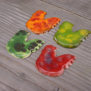 Op de afbeelding: Vier keramische haanfiguren in verschillende kleuren. De hanen zijn geschilderd in tinten groen, oranje, rood en geel.