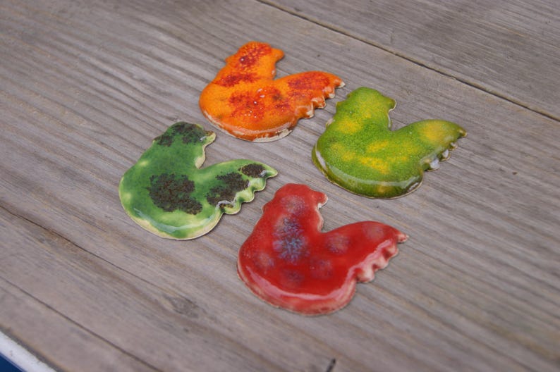 Op de afbeelding: Vier keramische haanfiguren in verschillende kleuren. De hanen zijn geschilderd in tinten groen, oranje, rood en geel.