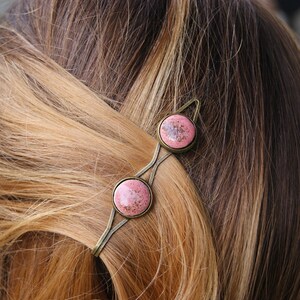 Puede incluir: Una pinza para el pelo dorada con dos piedras redondas de color rosa y blanco. La pinza est&aacute; en el pelo de una mujer.